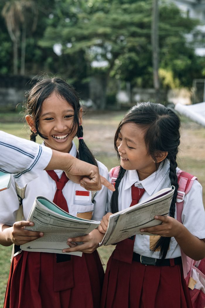 our-services-1 Two schoolgirls in uniforms smiling and reading book together outdoors.