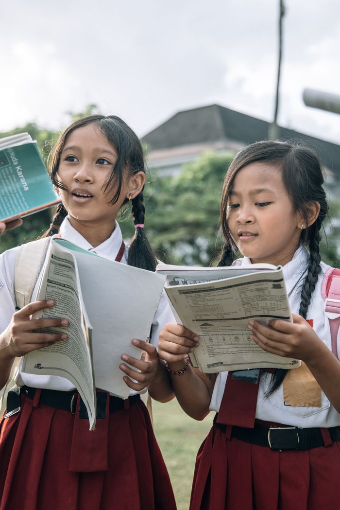 our-services-1 Two Asian girls in red school uniforms reading books outdoors, representing education and learning.
